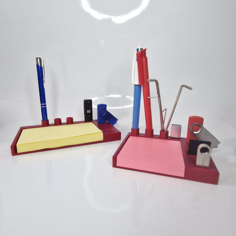 May include: Two red desk organizers with pens, sticky notes, and other office supplies. One has a blue pen and yellow sticky notes, the other has a red pen and pink sticky notes. Both have a small black item and a blue item.
