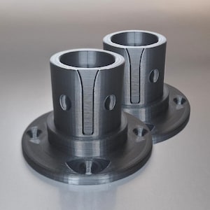 May include: Two gray metal cylindrical components with circular bases and central openings. Each component has a series of evenly spaced holes and vertical grooves. The components are designed for mechanical or industrial applications.