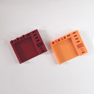 May include: Two plastic, square-shaped building blocks. One is a deep red, the other is orange. Each block has three circular holes and a series of vertical lines on one side. The blocks are on a white surface.