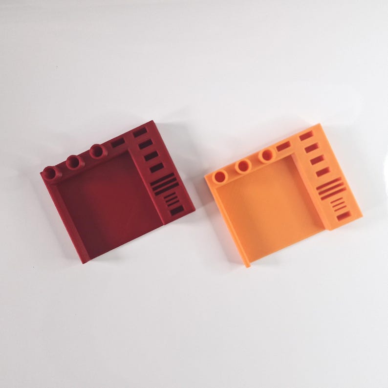 May include: Two plastic, square-shaped building blocks. One is a deep red, the other is orange. Each block has three circular holes and a series of vertical lines on one side. The blocks are on a white surface.
