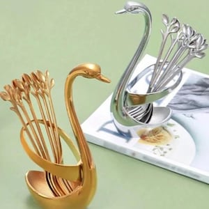 May include: Two swan-shaped holders, one gold and one silver, each containing small spoons. The spoons have a leaf-like design. The holders are on a light green surface, with a magazine visible in the background.