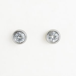 May include: A pair of silver-toned stud earrings, each featuring a single round, clear, faceted stone securely held within a smooth, polished metal bezel setting. The design is simple and circular, showcasing a bright, reflective finish.