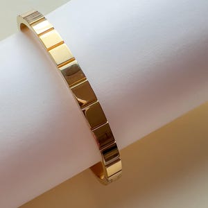 Könnte beinhalten: Ein goldfarbenes Armband mit einem strukturierten, rechteckigen Design.