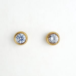 May include: A pair of round stud earrings featuring clear, faceted stones set in polished gold-tone metal bezels. The circular design highlights the brilliant cut of the stones, which are centered within the smooth, metallic frames for a classic look.