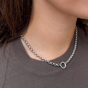 Puede incluir: Un collar de cadena plateada con eslabones redondos y un cierre circular, acompa&ntilde;ado de un pendiente de aro plateado a juego. El collar descansa sobre una camiseta sin mangas de color marr&oacute;n oscuro y el pendiente se lleva en el l&oacute;bulo.