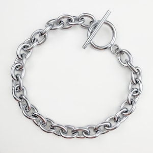 May include: A silver-colored chain bracelet with a toggle clasp. The bracelet features large, oval-shaped links. The clasp is a bar that fits through a circular loop. The jewelry is displayed on a white surface.