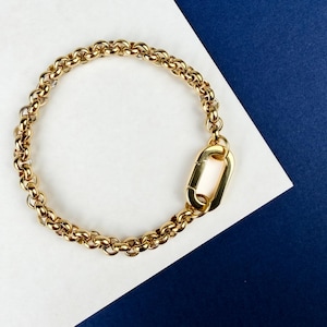 Peut inclure: Un bracelet doré avec un motif de chaîne et un grand fermoir rectangulaire. Le bracelet est présenté sur un fond blanc et bleu marine. Les maillons de la chaîne sont petits et rapprochés.