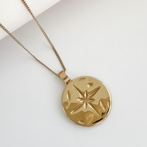 May include: Gold-colored necklace featuring a round pendant with a textured surface and a starburst design. The pendant hangs from a delicate chain. The necklace is displayed against a white background.