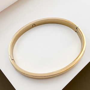 May include: A gold-coloured bangle bracelet with a hinged clasp. The bracelet has a smooth, polished surface and a simple, elegant design. The bracelet is approximately 6.35 cm in diameter.