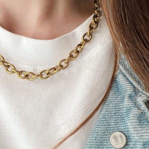 May include: A gold-colored chain necklace with large, oval links. The necklace is worn over a white t-shirt and a blue denim jacket. The chain is a bold statement piece.