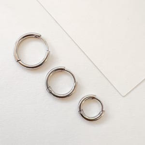 May include: Three silver-colored hoop earrings of varying sizes arranged on a white surface. The earrings are circular with a hinged clasp. The largest earring is at the top, followed by a medium and a small one.