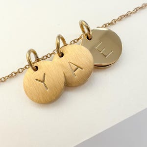 May include: A gold necklace featuring three circular pendants, each with a brushed metal texture and an engraved initial: Y, A, and E. The fine chain is also gold in colour.