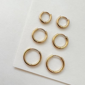 Peut inclure: Six boucles d'oreilles créoles dorées de différentes tailles sont disposées sur une surface blanche. Les boucles d'oreilles ont un design simple avec une fermeture à charnière. Le métal a une finition polie.