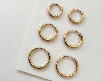 May include: Six gold-colored hoop earrings in varying sizes are arranged on a white surface. The earrings have a simple, classic design with a hinged closure. The metal has a polished finish, reflecting light.