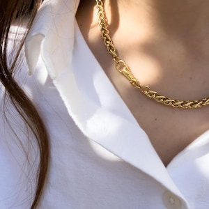 May include: A gold-toned wheat chain necklace featuring a polished finish and a secure lobster claw clasp. The chain consists of intricate, braided metal links that create a textured, rope-like appearance resting against a crisp white collared shirt.