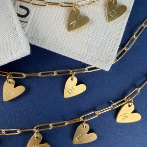 May include: Gold-colored chain necklaces with heart-shaped pendants. Each heart pendant has a single letter engraved on it. The necklaces are displayed on a white cloth against a dark blue background. The letters on the pendants are A, D, M, W, S, and L.
