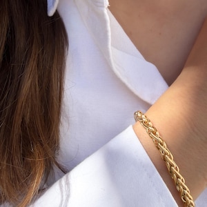 May include: A gold-toned bracelet featuring a thick, braided chain link design. The bracelet is constructed from polished metal links that interlock to create a continuous, textured rope-like appearance, resting against a crisp white fabric sleeve.