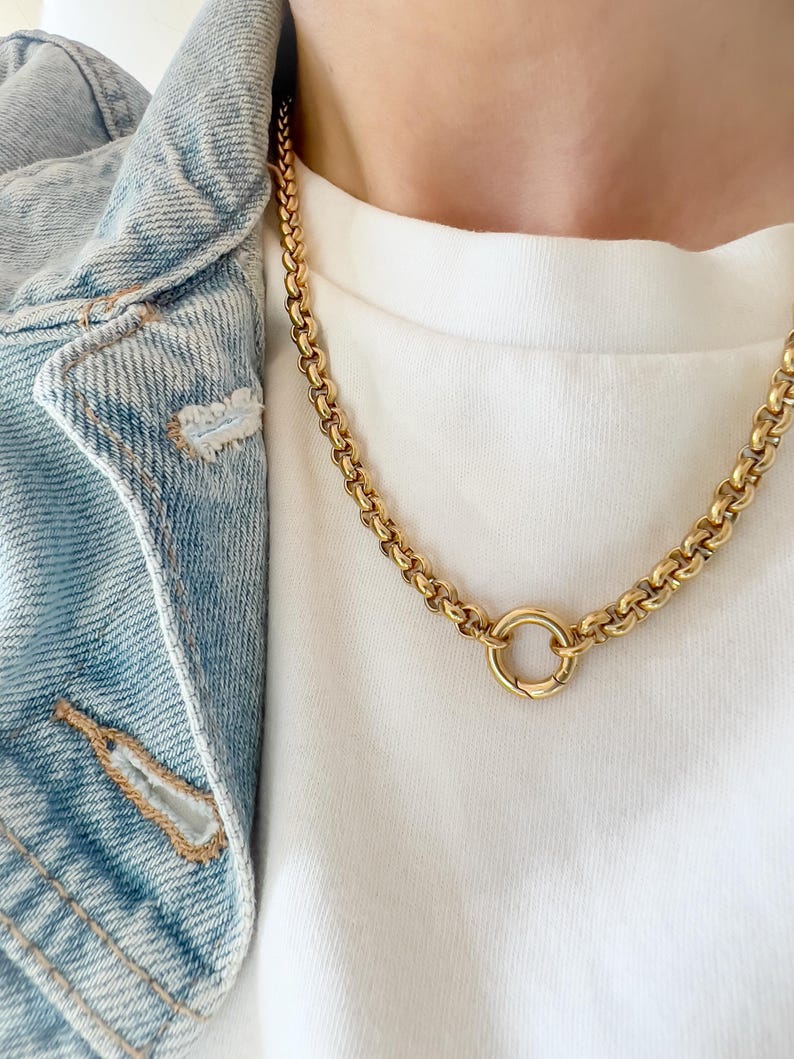 Puede incluir: Un collar de cadena en tono dorado con eslabones gruesos y redondeados, que presenta un prominente cierre de resorte circular en el centro. El collar se lleva sobre una camiseta blanca lisa y una chaqueta vaquera azul claro con costuras visibles.