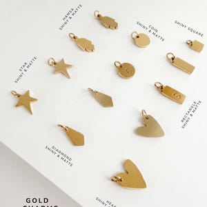 May include: A collection of gold-toned metal charms in various shapes including hamsa, coin, star, diamond, rectangle, square, and heart. Each charm features a small jump ring for attachment. Text labels identify the shapes and finishes as shiny and matte.