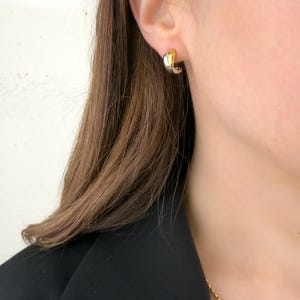 May include: A pair of two-tone gold and silver hoop earrings. The earrings are small, with a curved design. The earrings are worn on the ear of a person wearing a black blazer and a gold chain necklace.