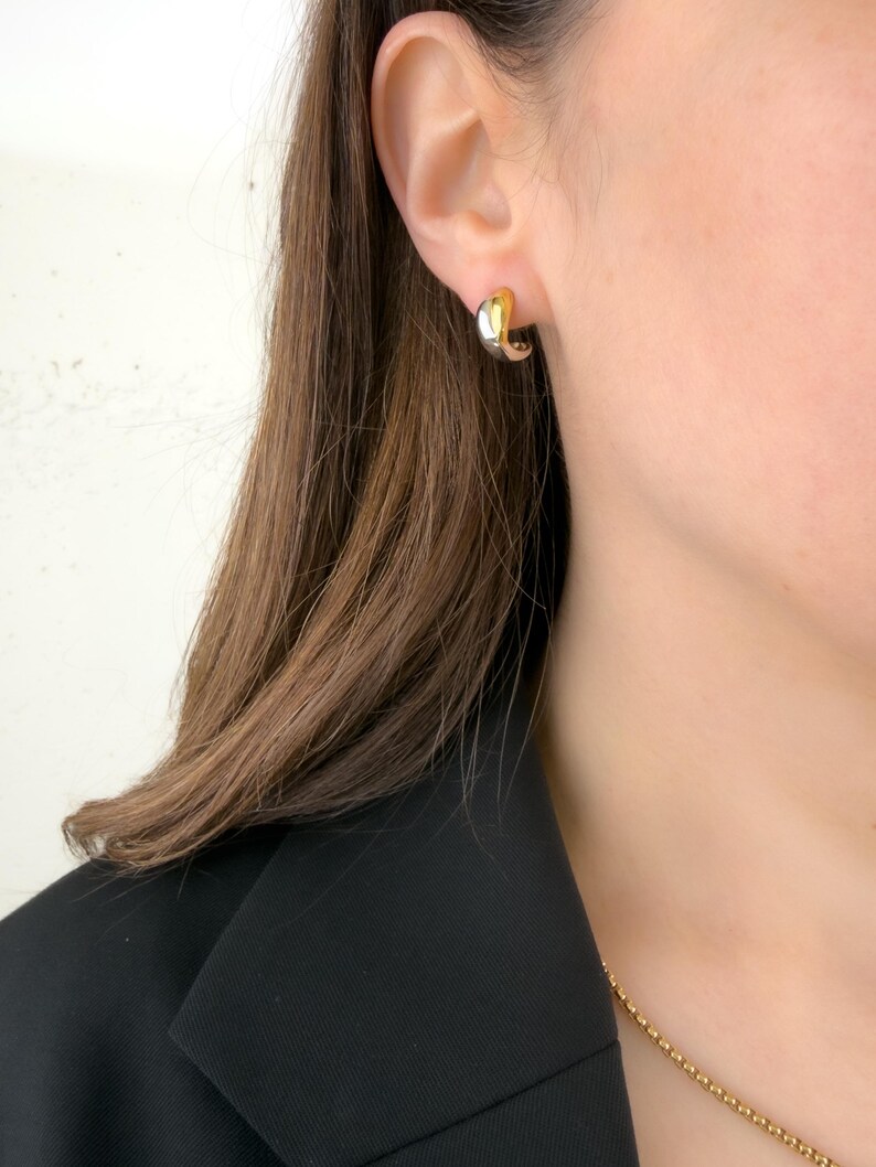 May include: A pair of two-tone gold and silver hoop earrings. The earrings are small, with a curved design. The earrings are worn on the ear of a person wearing a black blazer and a gold chain necklace.