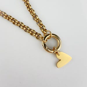 Pode incluir: Colar dourado com um design de corrente grossa. Inclui um fecho circular grande e um pingente em forma de coração. A joia é exibida em um fundo branco, destacando seu brilho metálico.
