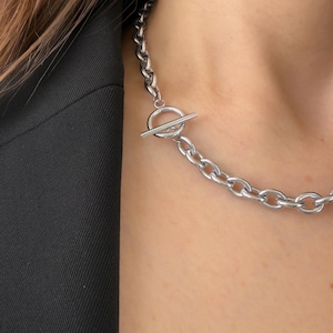 Puede incluir: Collar de cadena plateada con cierre de palanca. El collar presenta eslabones grandes y ovalados y un cierre sencillo de barra y anillo. La joya se lleva sobre una blazer negra.