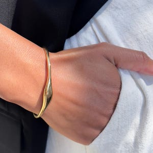 Puede incluir: Un brazalete dorado con un diseño suave y fluido. El brazalete tiene una forma curva y parece estar hecho de metal. Se aprecia en una muñeca, con una blazer negra y una camisa blanca en el fondo.