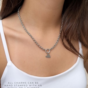 Puede incluir: Un collar de cadena de tono plateado con un cierre circular y un peque&ntilde;o colgante plano en forma de coraz&oacute;n. El texto en la imagen dice: ALL CHARMS CAN BE HAND STAMPED WITH AN INITIAL OR A NUMBER. El collar se lleva alrededor del cuello.