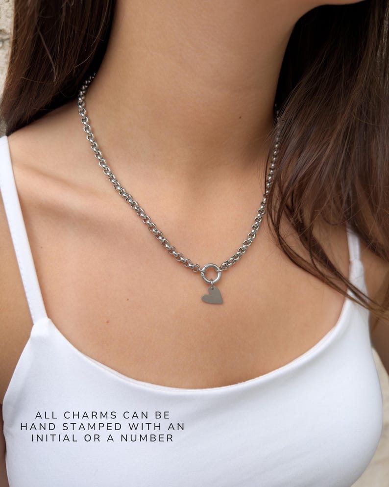 Puede incluir: Un collar de cadena de tono plateado con un cierre circular y un peque&ntilde;o colgante plano en forma de coraz&oacute;n. El texto en la imagen dice: ALL CHARMS CAN BE HAND STAMPED WITH AN INITIAL OR A NUMBER. El collar se lleva alrededor del cuello.