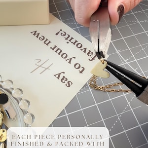 May include: A gold heart pendant necklace is being worked on with pliers. The necklace is on a gray surface with a white card that says "Say Hi to your new favorite!" and "Each piece personally finished & packed with care in SF."