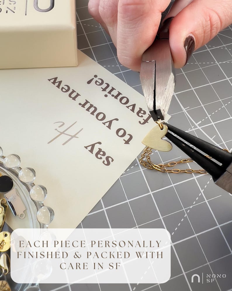 May include: A gold heart pendant necklace is being worked on with pliers. The necklace is on a gray surface with a white card that says "Say Hi to your new favorite!" and "Each piece personally finished & packed with care in SF."