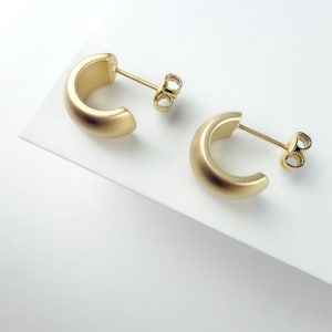 May include: A pair of gold-toned hoop earrings with a matte finish. These earrings feature a thick, semi-circular design and include standard post-back closures. The metal has a smooth, consistent texture throughout the entire surface of the hoops.