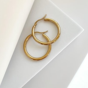 May include: A pair of gold hoop earrings. The earrings are circular and have a polished finish. They are displayed on a white surface with a folded paper element in the background. These earrings are a classic jewellery item.