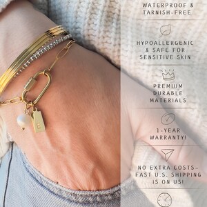 May include: A set of three gold-toned bracelets, including a smooth bangle, a beaded chain, and a link chain with a carabiner clasp. The carabiner features a small white pearl charm and a rectangular gold tag engraved with the letter E.