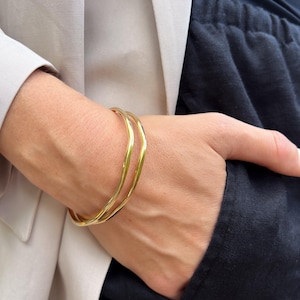 Puede incluir: Un brazalete dorado con un dise&ntilde;o ondulado. El brazalete se lleva en la mu&ntilde;eca, con una blazer de color neutro y pantalones oscuros en el fondo. El brazalete parece ser de metal.