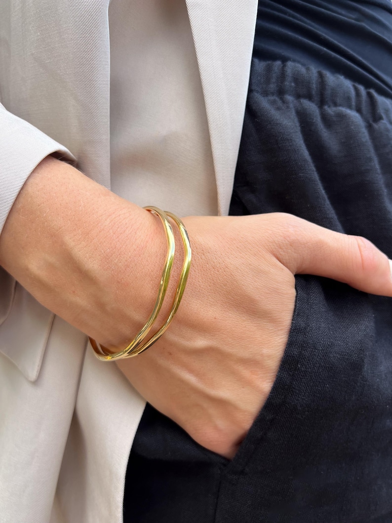 Puede incluir: Un brazalete dorado con un dise&ntilde;o ondulado. El brazalete se lleva en la mu&ntilde;eca, con una blazer de color neutro y pantalones oscuros en el fondo. El brazalete parece ser de metal.