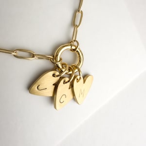 May include: A gold-toned necklace featuring a thick paperclip chain and a circular clasp. Three heart-shaped charms are attached to the clasp, each engraved with a single letter: J, C, and M. The metal has a brushed, matte finish throughout.