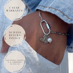 May include: A silver-toned chain bracelet featuring a carabiner-style clasp, a heart-shaped charm, a small rectangular tag engraved with the letter A, and a single white pearl bead. The bracelet is worn against a light-coloured fabric background.