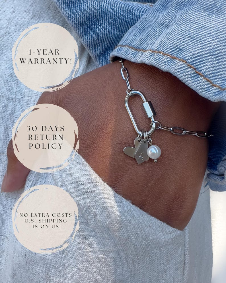 May include: A silver-toned chain bracelet featuring a carabiner-style clasp, a heart-shaped charm, a small rectangular tag engraved with the letter A, and a single white pearl bead. The bracelet is worn against a light-coloured fabric background.