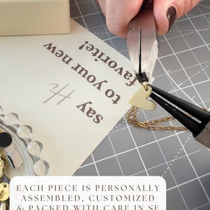 Puede incluir: Un dije de coraz&oacute;n dorado se sujeta a una cadena dorada con alicates de metal. Una tarjeta con el texto 'say Hi to your new favorite!' y 'EACH PIECE IS PERSONALLY ASSEMBLED, CUSTOMIZED & PACKED WITH CARE IN SF' se ve sobre una alfombrilla cuadriculada.