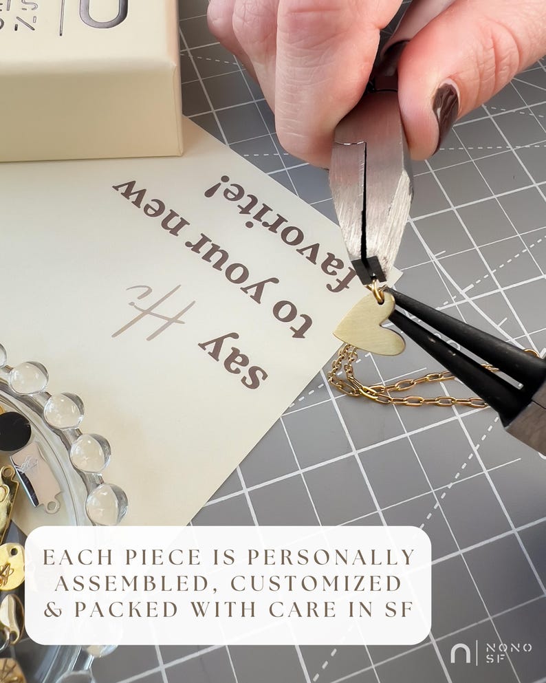 Puede incluir: Un dije de coraz&oacute;n dorado se sujeta a una cadena dorada con alicates de metal. Una tarjeta con el texto 'say Hi to your new favorite!' y 'EACH PIECE IS PERSONALLY ASSEMBLED, CUSTOMIZED & PACKED WITH CARE IN SF' se ve sobre una alfombrilla cuadriculada.