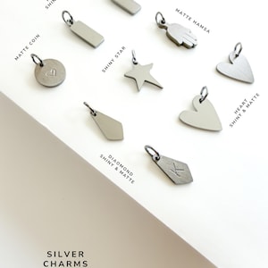 May include: A collection of silver charms including a matte coin with a heart, a shiny star, a matte hamsa, a diamond shape with the letter K, and various rectangle and heart shapes labelled as shiny and matte silver charms options.