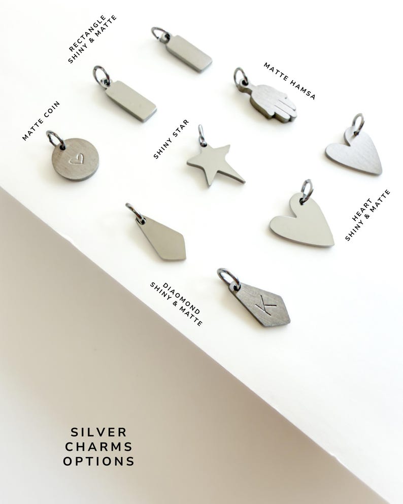 May include: A collection of silver charms including a matte coin with a heart, a shiny star, a matte hamsa, a diamond shape with the letter K, and various rectangle and heart shapes labelled as shiny and matte silver charms options.