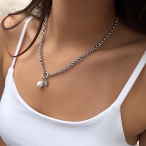 Puede incluir: Un collar de cadena plateada con un dije de perla blanca redonda y un peque&ntilde;o colgante plano en forma de coraz&oacute;n plateado, unidos a un anillo circular. El collar descansa sobre una camiseta de tirantes blanca lisa.
