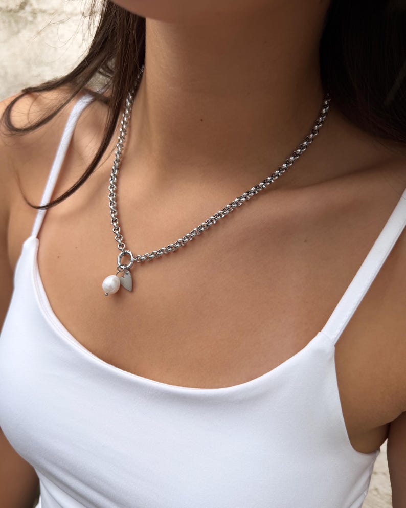 Puede incluir: Un collar de cadena plateada con un dije de perla blanca redonda y un peque&ntilde;o colgante plano en forma de coraz&oacute;n plateado, unidos a un anillo circular. El collar descansa sobre una camiseta de tirantes blanca lisa.