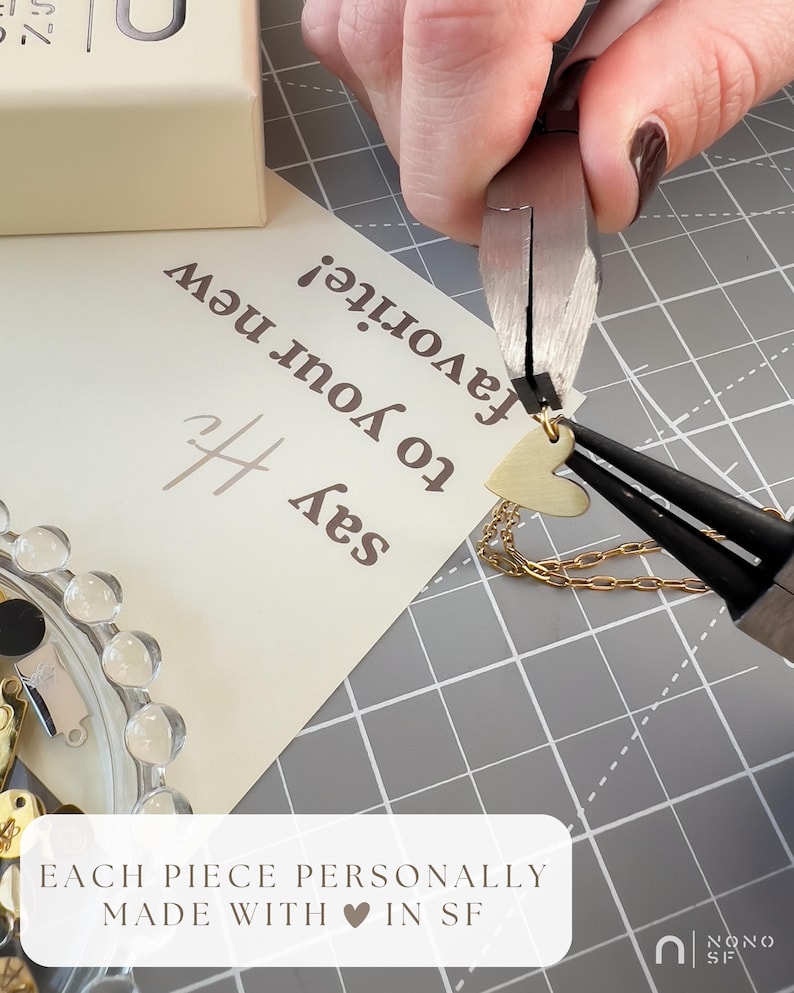 May include: A gold-toned heart charm being attached to a gold chain with metal pliers. A cream-coloured card with the text 'say Hi to your new favourite!' and a small box are visible. A white label reads 'EACH PIECE PERSONALLY MADE WITH HEART IN SF'.
