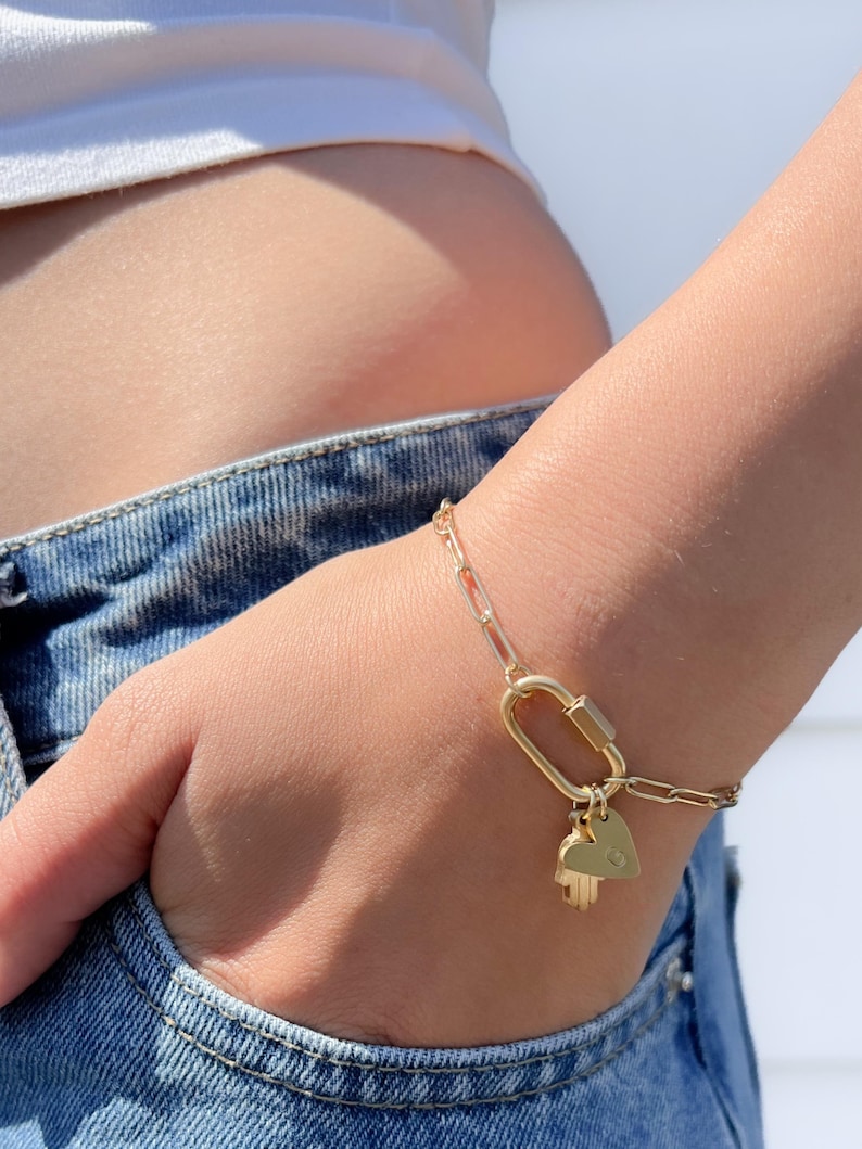 May include: A gold-toned chain bracelet featuring elongated oval links and a central carabiner-style clasp. Two small gold-toned charms are attached to the clasp: one shaped like a heart and the other shaped like a hamsa hand symbol.