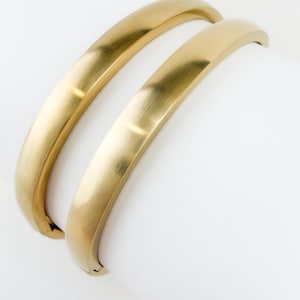 May include: Two gold-toned metal bangle bracelets with a smooth, brushed matte finish. Each bracelet features a curved, semi-circular profile and a hinged closure mechanism, displaying a consistent metallic sheen across the surface.