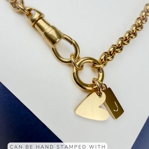 May include: A gold-toned chain necklace featuring a swivel clasp, a circular ring, and two charms: one heart-shaped and one rectangular with the letter J. The text CAN BE HAND STAMPED WITH AN INITIAL OR A NUMBER is visible at the bottom.